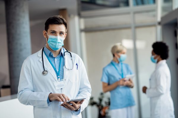 Doctor with protective face mask using touchpad while standing in hallway at medical clinic and looking at camera.