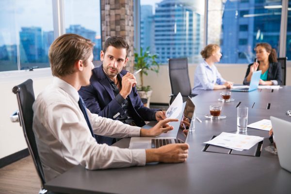 Two serious young businessmen sitting in office, discussing business issues and two businesswomen working on background