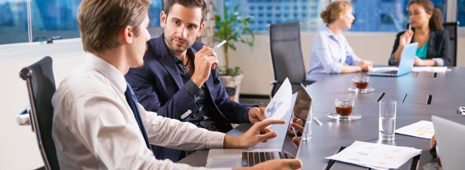 Two serious young businessmen sitting in office, discussing business issues and two businesswomen working on background